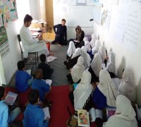 Young children listen to their teacher in a narrow classroom. All are seated on the floor
